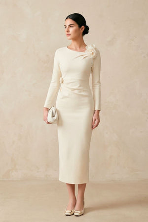 A full-length view of an ivory fitted midi dress featuring long sleeves, a subtle waist detail, and floral embellishment at the shoulder, styled with neutral heels and a clutch.
