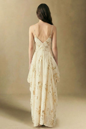Model seen from the back wearing an ivory maxi dress with delicate gold sequin floral details, thin straps, and a softly draped silhouette against a warm studio backdrop.
