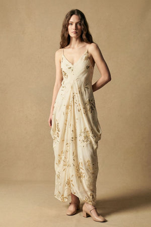 Model wearing an ivory maxi dress with subtle gold sequin floral motifs, a V-neckline, and fluid drape, styled with neutral heels in a softly lit studio setting.