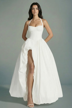 Model wears an ivory halter corset maxi dress with a softly gathered bodice, cinched waist, and dramatic front slit, styled with minimal heels against a clean studio background.