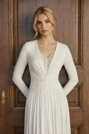 A model stands against a carved wooden door wearing an ivory long sleeve maxi dress with a lace V-neck and gently cinched waist.