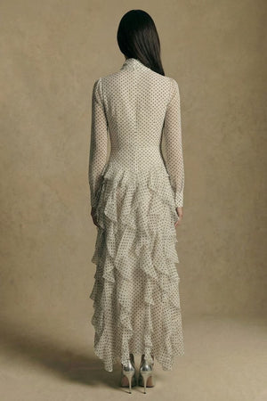 Back view of a model wearing an ivory polka dot maxi dress with a clean fitted back, long sheer sleeves, and a tiered ruffle skirt, photographed in a warm neutral studio environment.