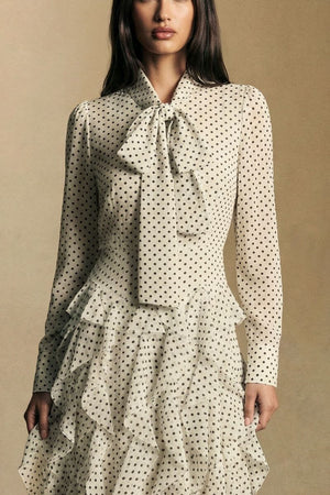 Close-up of the bodice of an ivory polka dot maxi dress highlighting the soft bow-tie neckline, sheer long sleeves, and textured ruffle detailing at the waist.