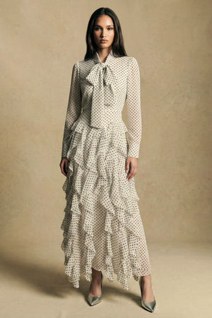 A model wearing an ivory polka dot long-sleeve maxi dress with a bow-tie neckline and cascading ruffle skirt, styled with pointed metallic heels against a warm neutral studio backdrop.