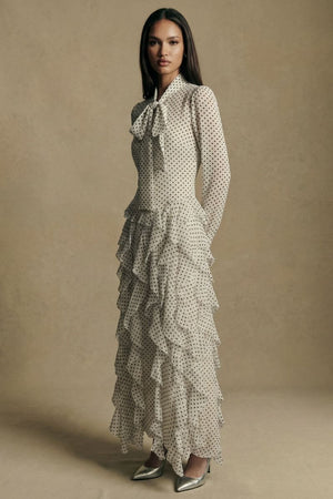Side profile of a model showcasing the slim silhouette and flowing ruffle layers of an ivory polka dot long-sleeve maxi dress, paired with metallic pointed heels.