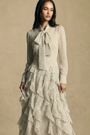 Three-quarter view of a model wearing an ivory polka dot maxi dress with sheer long sleeves, delicate ruffles, and a feminine bow neckline, styled in an elegant neutral-tone studio.