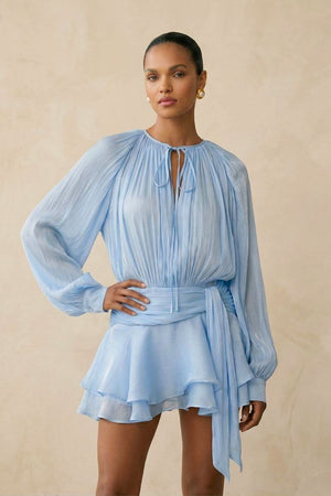 Model posed slightly angled in a light blue chiffon mini dress featuring airy pleats, a soft tie neckline, gathered waist, and flowing ruffle hem for a feminine silhouette.