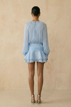 Model shown from the back wearing a light blue chiffon mini dress with soft pleated long sleeves, a gathered waist sash, and a layered ruffle hem against a warm neutral studio background.