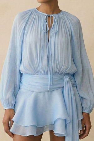 Close-up of the light blue chiffon mini dress highlighting the softly pleated bodice, tie neckline detail, sheer long sleeves, and fluid texture of the fabric.