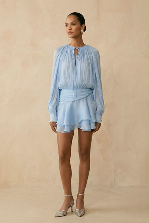 Model wearing a light blue chiffon mini dress with a relaxed blouson top, cinched waist sash, long sheer sleeves, and layered ruffle skirt, styled with delicate heels in a neutral studio setting.