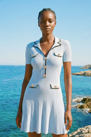 Close-up of a woman in a light blue knit mini dress near the ocean, showing pearl buttons and contrast trim.