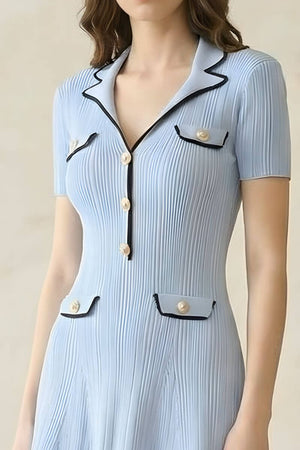 A woman wearing a light blue short-sleeve knit mini dress with pleated texture, contrast black piping, and pearl buttons on the collar and pockets.