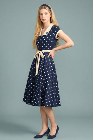A side view of a model wearing a navy polka dot midi dress with an ivory collar and waist tie, showing the fluid skirt movement and classic feminine silhouette.