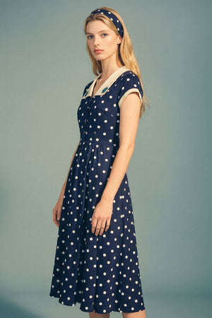 A model poses in a navy polka dot midi dress with an ivory collar and button-front detail, styled with a matching headband for a refined vintage-inspired look.
