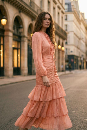 A model walks along a quiet European street wearing a peach chiffon maxi dress with long sheer sleeves, a softly ruched bodice, delicate front ruffle detailing, and a flowing tiered skirt that moves lightly with each step in warm evening light.