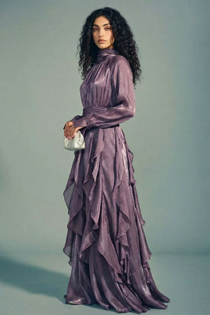 Model standing in a side pose wearing a plum satin maxi dress with a high gathered neckline, sheer long sleeves, and cascading ruffle layers, styled with a small metallic clutch.
