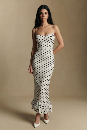 A model stands against a neutral backdrop wearing an ivory polka dot midi dress with a fitted silhouette, subtle mermaid shape, and ruffled hem, paired with white pointed heels.