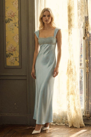 A model wears a powder blue satin maxi dress with a beaded neckline, styled with ivory heels and a matching handbag in a softly lit vintage interior.