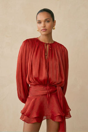 A close view of the red chiffon mini dress highlighting the pleated neckline with a delicate tie detail, sheer long sleeves, and fluid texture that drapes softly over the body.