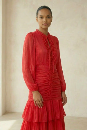 A model wears a red chiffon tiered midi dress with long sleeves, a ruffled neckline and ruched bodice, standing in soft natural studio lighting.