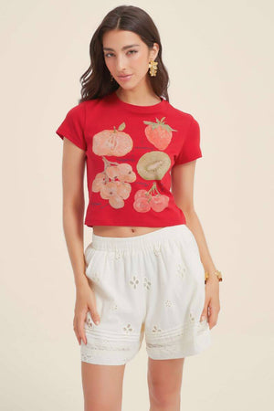 Front view of a woman wearing a red cropped T-shirt with illustrated fruit print paired with white eyelet lace shorts, styled with gold floral earrings and neutral heels.
