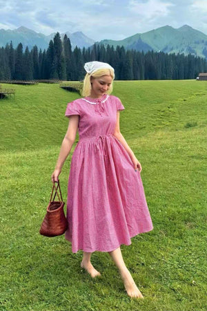 Woman wearing a red gingham midi dress with lace collar and tie-waist styling in a grassy, mountainous outdoor setting, carrying a woven bag.