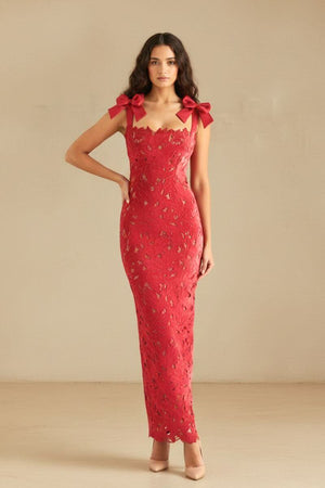 Full-length view of a woman wearing a red lace bow-shoulder maxi dress with a fitted silhouette and nude heels on a neutral background.