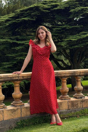 Woman in a red ruffle polka dot maxi dress posing gracefully on a stone terrace surrounded by lush green trees, styled with red flats for a vintage outdoor look.