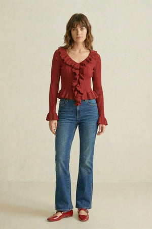 Woman in a red ribbed knit ruffle top paired with blue straight-leg jeans and red flats standing in a neutral studio setting.