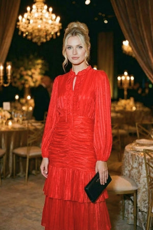 A woman in a red tiered chiffon midi dress with a ruffled neckline poses at an elegant evening event surrounded by chandeliers and candlelit tables.