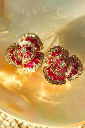 Ruby and crystal floral stud earrings in gold setting, styled on a reflective surface with warm golden light and soft shadows.