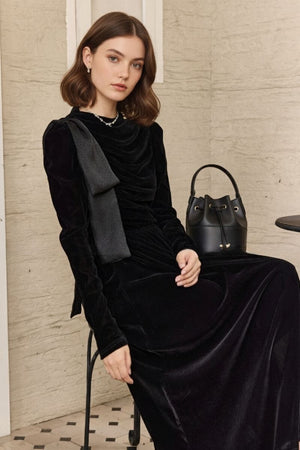 Model seated by a small café table wearing a black velvet maxi dress with a softly ruched bodice and shoulder bow, accessorized with a pearl necklace; black bucket bag rests in the background.