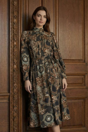 A woman stands against carved wooden doors wearing a long-sleeve midi dress with a high neckline, gathered waist, and vintage-inspired floral and medallion print.