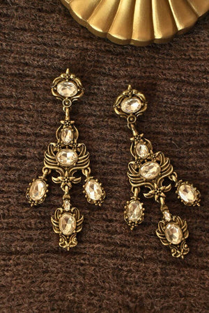 Vintage-inspired gold drop earrings featuring layered clear crystals and intricate baroque-style metalwork, photographed on a brown textile backdrop.