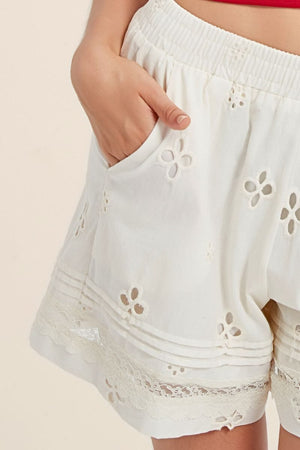 Close-up of white embroidered eyelet shorts showing pocket stitching, floral cut-outs, and lace trim at the hem for a soft summer look.