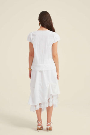Back view of a woman wearing a white lace-trim blouse with puff sleeves and a peplum hem, styled with a white layered lace midi skirt.