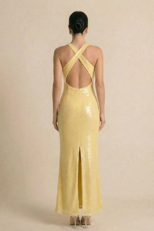 Back view of a yellow sequin evening gown showcasing cross-back straps, an open back, and a center back slit, modeled in a neutral-toned studio.