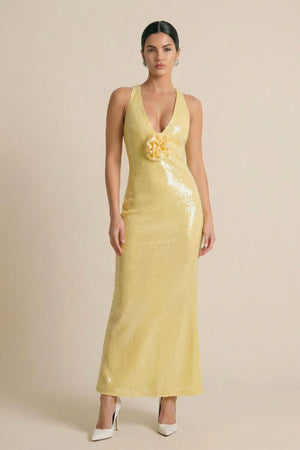 Full front view of a model wearing a yellow sequin deep-V maxi gown with a floral appliqué at the bust, styled with white heels in a minimal beige studio.