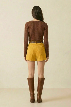 Back view of a woman wearing a brown ribbed long-sleeve sweater paired with high-waisted yellow corduroy shorts and tall brown boots.