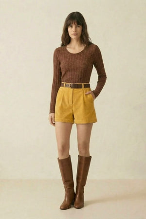 A woman in a brown ribbed scoop-neck sweater and yellow corduroy shorts styled with a brown belt and tall brown boots, standing in a clean studio setting.