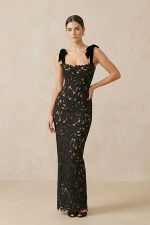 model wearing a full-length black lace gown with velvet bow straps and fitted silhouette, standing in a neutral beige studio.