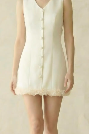 model wearing an ivory sleeveless button-front mini dress with soft rosette trim at the hem, standing against a neutral beige backdrop.