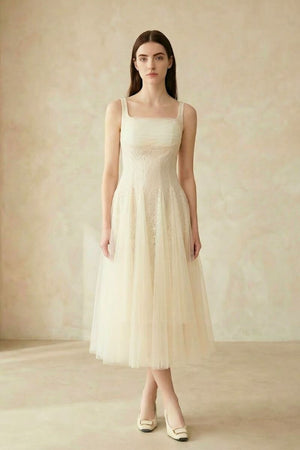 A woman wearing an ivory lace tulle midi dress with wide straps and a soft A-line silhouette, shown standing against a neutral backdrop.