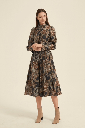 Model wearing a vintage-inspired high-neck midi dress with a baroque floral print in deep earthy tones, long puff sleeves, button-up bodice, and waist tie, paired with beige ankle boots.