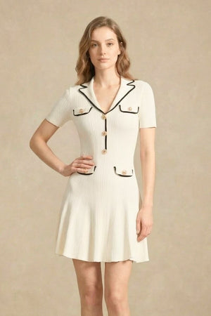 A close-up front view of a woman wearing a white mini dress with black contrast trim and decorative gold button pockets.
