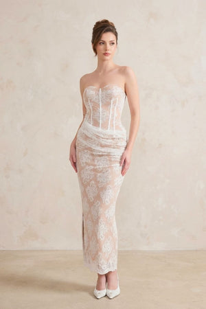A model in a strapless white lace corset maxi dress with a nude underlay stands in a studio, showcasing the draped hip detail and floral lace pattern.