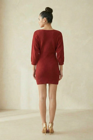 A woman faces away from the camera wearing a wine-red knit mini dress with three-quarter sleeves, shown in a clean studio setting to highlight the simple back view and fitted silhouette.