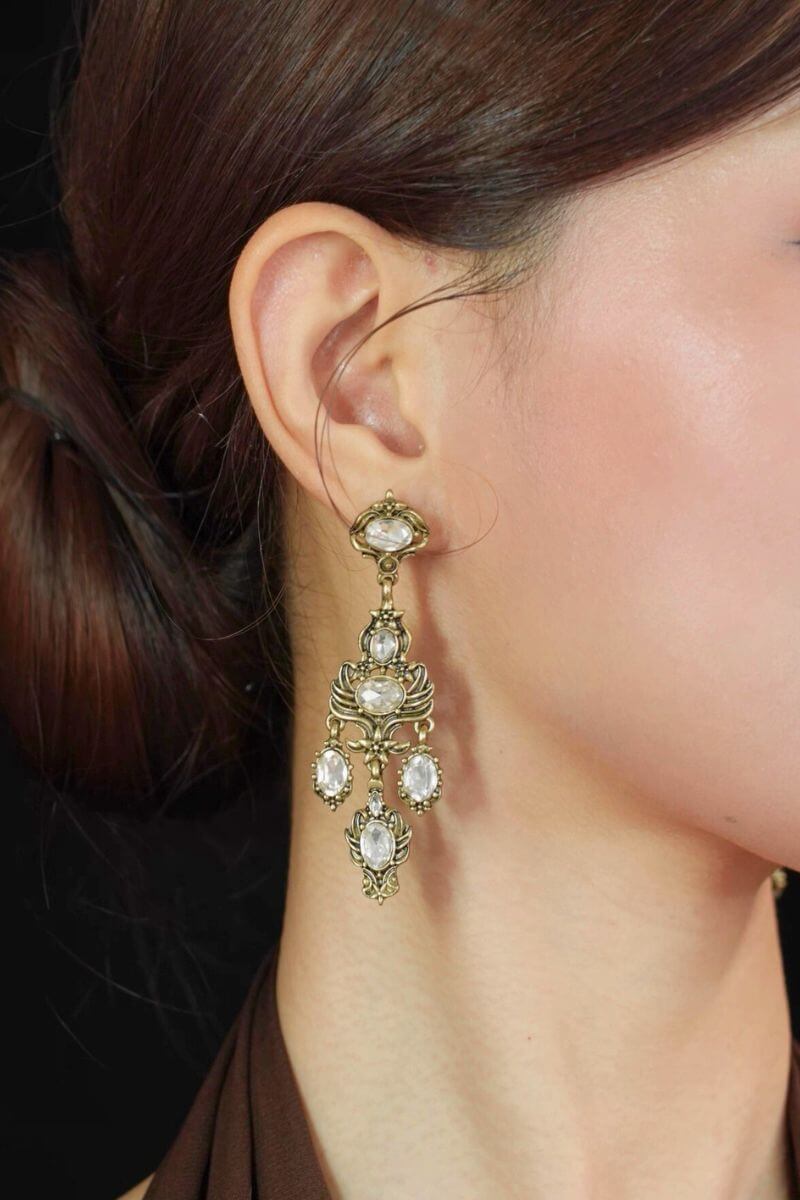 Model wearing antique gold chandelier earrings with clear faceted crystals and ornate vintage detailing, styled against a dark background.