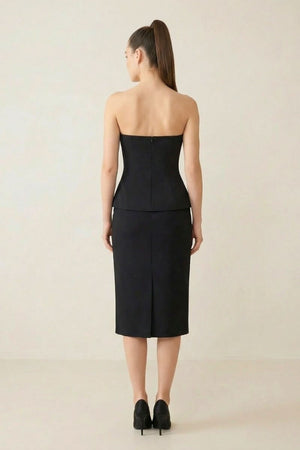 Rear view of a model in a black strapless peplum top with a center zipper and a matching black midi skirt.