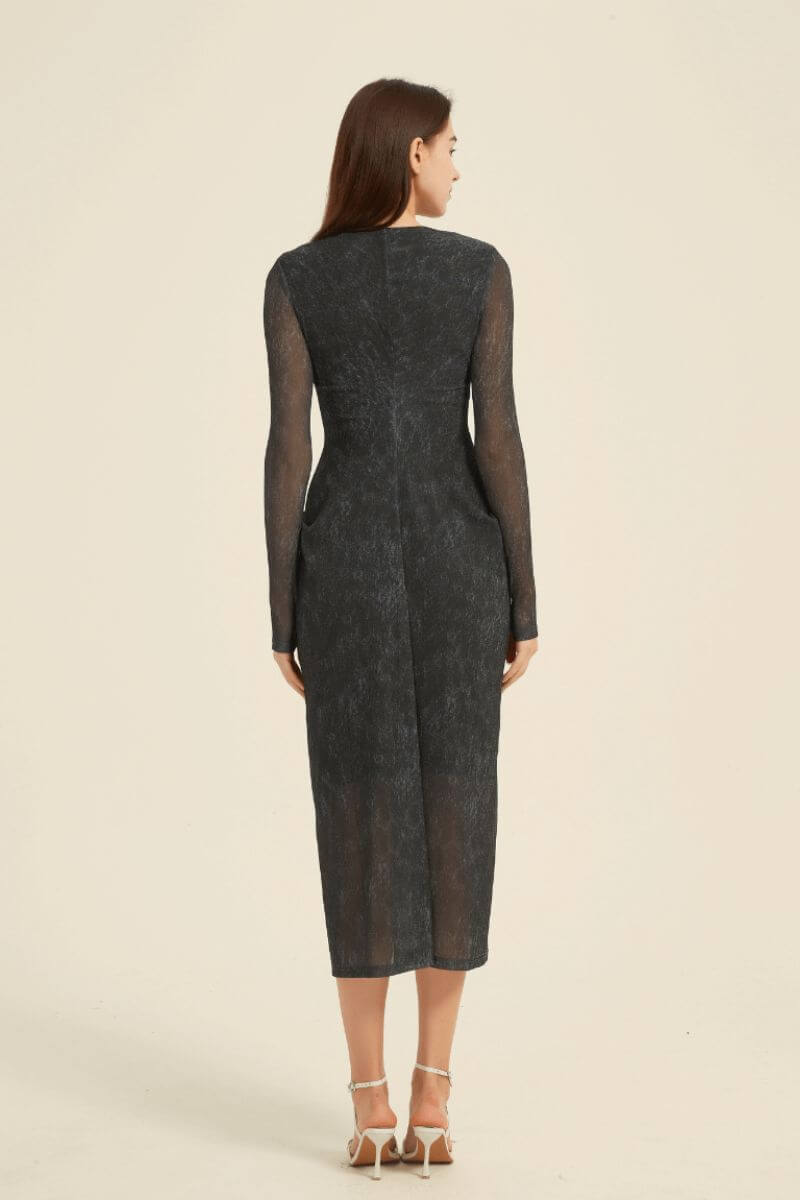 Rear view of a women’s black midi dress featuring sheer long sleeves, a V-neckline, and soft front pleats with a semi-sheer hem.